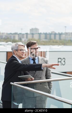 Mature businesswoman in eyeglasses pointing at something and discussing it with her colleague while they standing in the city Stock Photo
