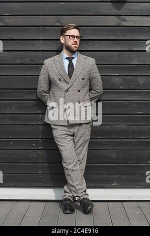 Young businessman in eyeglasses and in suit leaning on the wall in the city and looking away with pensive sight Stock Photo