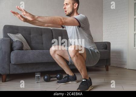 Young ordinary man go in for sport at home. Real picture of regular guy doing squats with stretch hands forward. Beginner or amateur has workout in Stock Photo