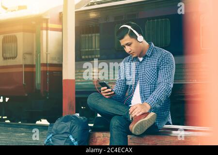 Asian young man using smartphone and listen music waiting train at train station. Stock Photo
