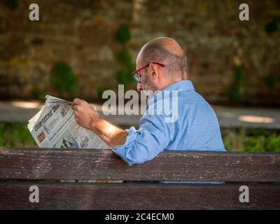 Rear view of caucasian middle aged man sitting on a bench reading a newspaper. Siena, Italy. Stock Photo