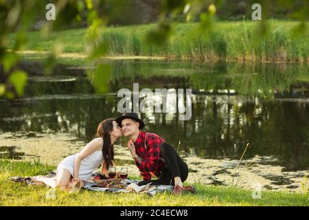 Kissing. Caucasian young, happy couple enjoying weekend together in the park on summer day. Look lovely, happy, cheerful. Concept of love, relationship, wellness, lifestyle. Sincere emotions. Stock Photo