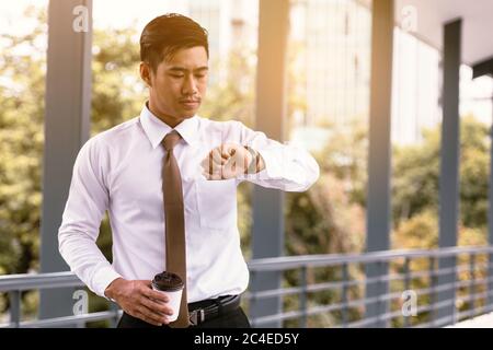 Young asian male standing checking time and holding coffee cup with confident looking at watches on hand at outside company. Stock Photo