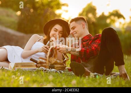 Clinking glasses with wine. Caucasian young couple enjoying weekend in the park on summer day. Look lovely, happy, cheerful. Concept of love, relationship, wellness, lifestyle. Sincere emotions. Stock Photo