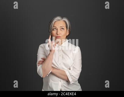 Thoughtful graying woman with forefinger pointing up, while looking up at empty place for text. Cut out on gray. Solution Idea concept Stock Photo