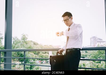 Young asian male standing checking time and holding briefcase with confident looking at watches on hand at outside company. Stock Photo