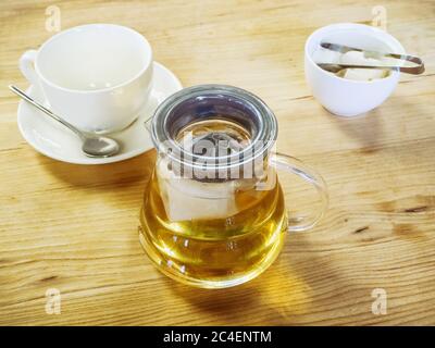 A transparent glass teapot with tea next to a vase with sugar and a clean cup stand on a wooden surface. Tea party Stock Photo