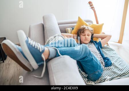 Sleeping preteen boy lying at the home living room filled with sunlight on the cozy sofa dressed casual jeans and sneakers listening to music Stock Photo
