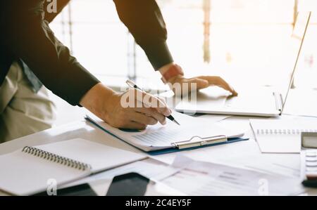 Businessman holding pen and pointing summary chart and analysis annual cost report with using laptop at room office desk. Stock Photo