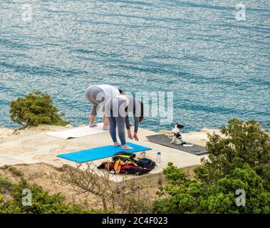 Two plump Woman in white and grey costume doing Yoga and looking at the dog on the beach near the sea. dog sitting on grey yoga mat. Copy space. The Stock Photo