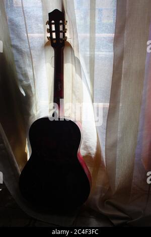silhouette of an old classical guitar seen from behind against the curtains of a window in the dark Stock Photo