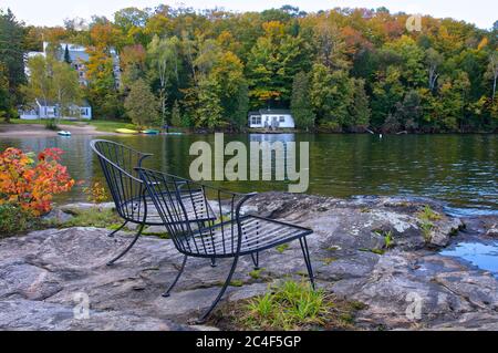 Retirement Living - Two outdoor chairs sitting on a rocky shore facing a calm lake with trees and a white Stock Photo
