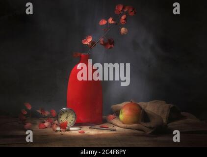 Classic still-life with a red vase, an apple, autumn leaves and  an alarm clock. Art photography. Stock Photo