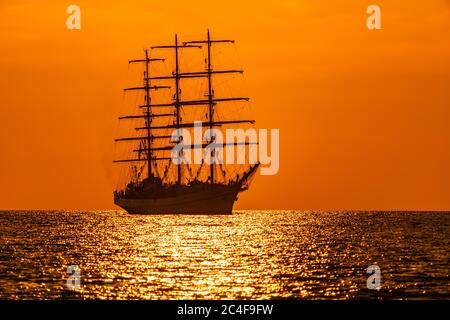 A red burning sunset with the silhouette of sailing ship. Sailors set sails on masts of ship. White sailing vessel floating in the sea. Copy space. Stock Photo