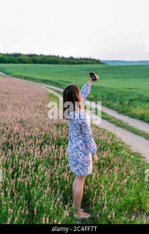 Young girl make selfie in a field of Pink flowers Sainfoin, Onobrychis viciifolia. The concept of an travel, relax, active and healthy life in harmony Stock Photo