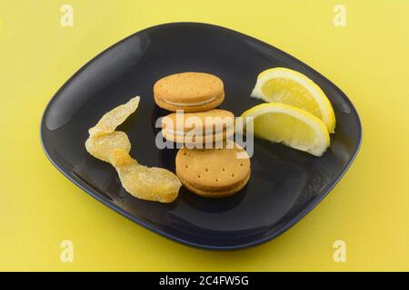 Cookies with lemon ginger cream filling with lemon wedges and crystallized ginger on dark blue snack plate on yellow background Stock Photo