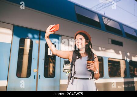 Theme travel, travel by rail. Beautiful caucasian woman in dress and backpack uses phone near railway carriage at station. A tourist takes selfie at