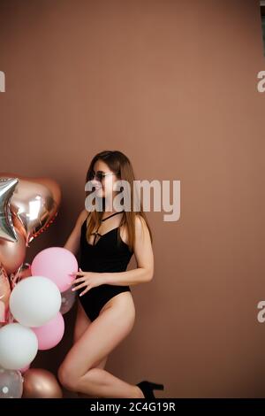 Slender girl in a bodysuit and colored balloons in her hands stands on a brown background in a festive mood posing in front of the camera. Stock Photo