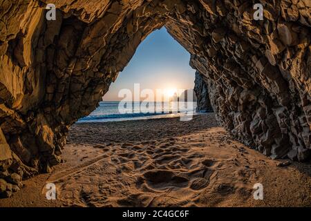 view from the stone cave on the sunset sea and the beach, the volcanic rock of the cave is lit by the warm setting sun Stock Photo