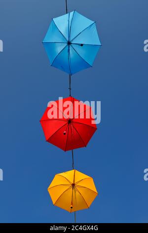Low angle view of three different colored umbrellas red, orange and blue suspended on a cable against a clear blue sky. Stock Photo