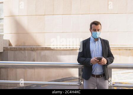 Man with a mask on his face and wearing a suit using his cell phone while leaning on a metal railing in the street Stock Photo