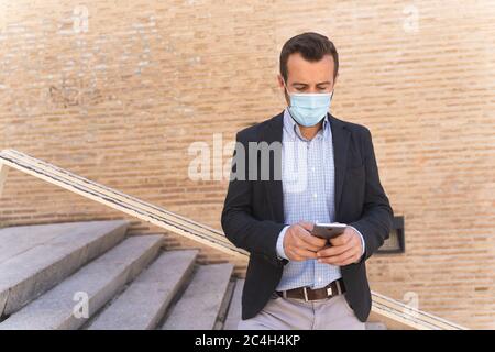 Businessman with a blue jacket and a mask on his face leaning against a railing while using his mobile Stock Photo