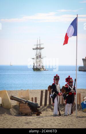 Malaga, Spain - October 26, 2014: Napoleonic soldiers of the Great Army next to a waving french flag and a cannon on a beach. A brig sails away. Histo Stock Photo