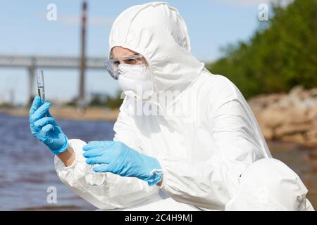 Scientist in protective workwear holding flask with liquid in her hands and examining it outdoors Stock Photo