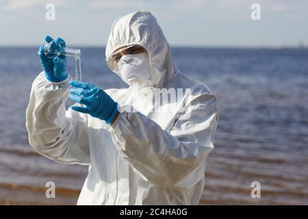 Scientist in protective suit pouring liquid from one flask to another while standing near the sea outdoors Stock Photo