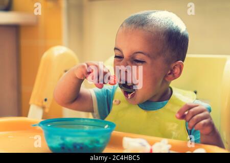 The boy 2 years eats porridge. Children's table. The concept of the child's independence. funny kid in a baby seat. toned Stock Photo