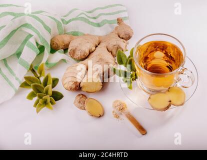 A glass cup of ginger tea with a fresh ginger root next to it and some green leaves around it, there is a white and green fabric at the corner. Stock Photo