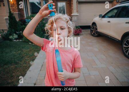 Young Caucasian girl blowing soap bubbles on home front yard. Child having fun outdoor on sunset. Authentic happy childhood magic moment. Stock Photo