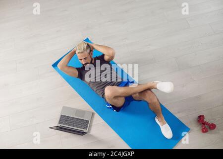 A young fit man does workout exercises on the abs while lying on the floor, watches an online trainer course while having a laptop in room. Stock Photo