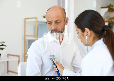 Young female cardiologist listening to senior man's heartbeat at clinic. Doctor examining elderly patient at office Stock Photo