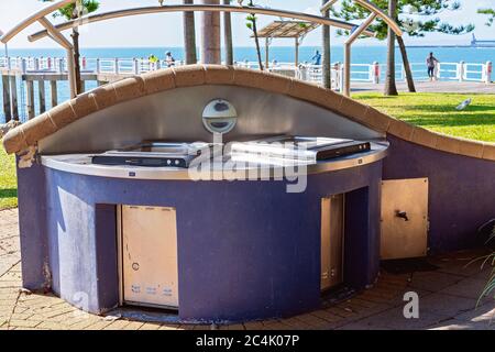 Public barbecue at the beach with people enjoying the sunshine on a recreational jetty in the background Stock Photo