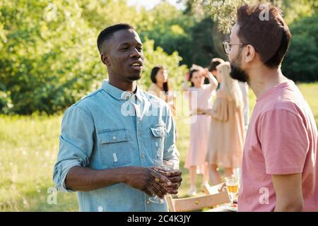 Young African man with glass of wine talking to mixed-race guy outdoors on background of their girlfriends having conversation Stock Photo