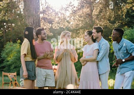 Company of three happy young couples in casualwear discussing news or their plans for summer while gathered together in park Stock Photo