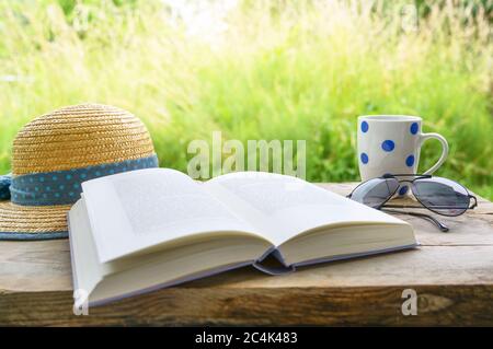 Holidays at home with book, sun glasses, coffee mug and straw hat on a rustic wooden table in the garden, safe relaxation during the coronavirus pande Stock Photo