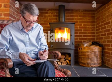 Senior caucasian adult checking a document with fountain pen in hand while seated. He is sitting by wood burning furnace Stock Photo