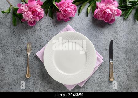 Festive table setting with pink peony, white dishware, silverware on gray. Top view. Stock Photo