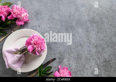 Festive table setting with pink peony, white dishware, silverware on gray. Top view. Stock Photo