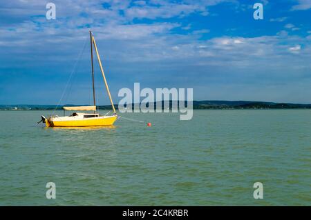 A yellow sailboat anchored on the water in slightly cloudy weather with mountains in the distance. Lake Balaton Stock Photo