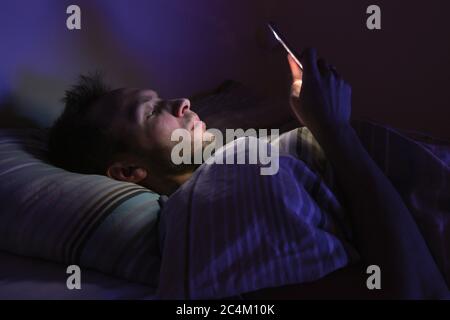 Closeup portrait of young sleepy exhausted man lying in bed using smartphone at late night, can not sleep.Insomnia, nomophobia, sleep disorder Stock Photo