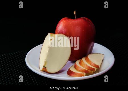 An apple, a quarter of an apple and apple slices all on a white plate and black background Stock Photo