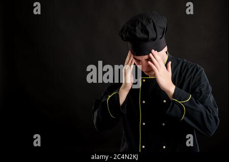 Portrait of young male dressed in a black chef suit with open arms looking up, has headache posing on a black isolated background with copy space adve Stock Photo