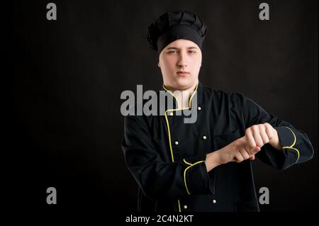 Portrait of young male dressed in a black chef suit showing to late gesture and looking her watch posing on a black isolated background with copy spac Stock Photo