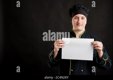 Portrait of young male dressed in a black chef suit holding a white paper and smileing posing on a black isolated background with copy space advertisi Stock Photo