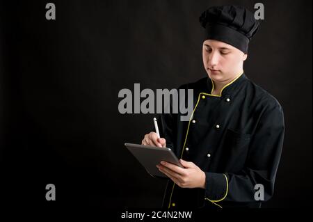 Portrait of young male dressed in a black chef suit takes notes, posing on a black isolated background with copy space advertising area Stock Photo