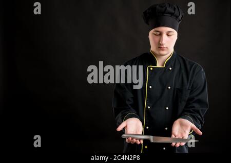 Young male dressed in a black chef suit showing knife posing on a black isolated background with copy space advertising area Stock Photo