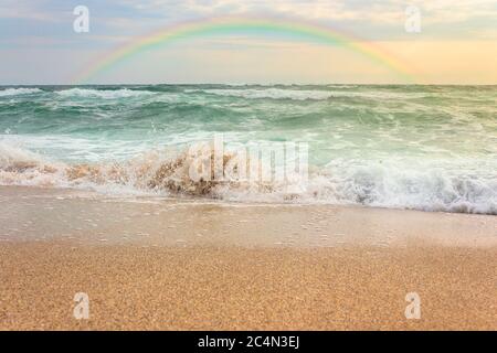storm on the sandy beach at sunset. dramatic ocean scenery with cloudy sky. rainbow above the rough water and crashing waves in evening light Stock Photo